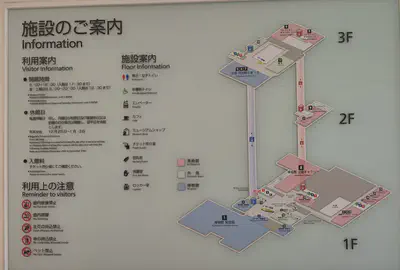 Floor directory at the Okinawa Prefectural Museum and Art Museum, Naha. The right half shows floors 1F, 2F and 3F drawn in perspective and stacked vertically, connected by lift shafts. A red “You are here” marker sits on the ground floor near the sub-entrance. Galleries are numbered. The left half gives opening hours and visitor rules. There is no north arrow.